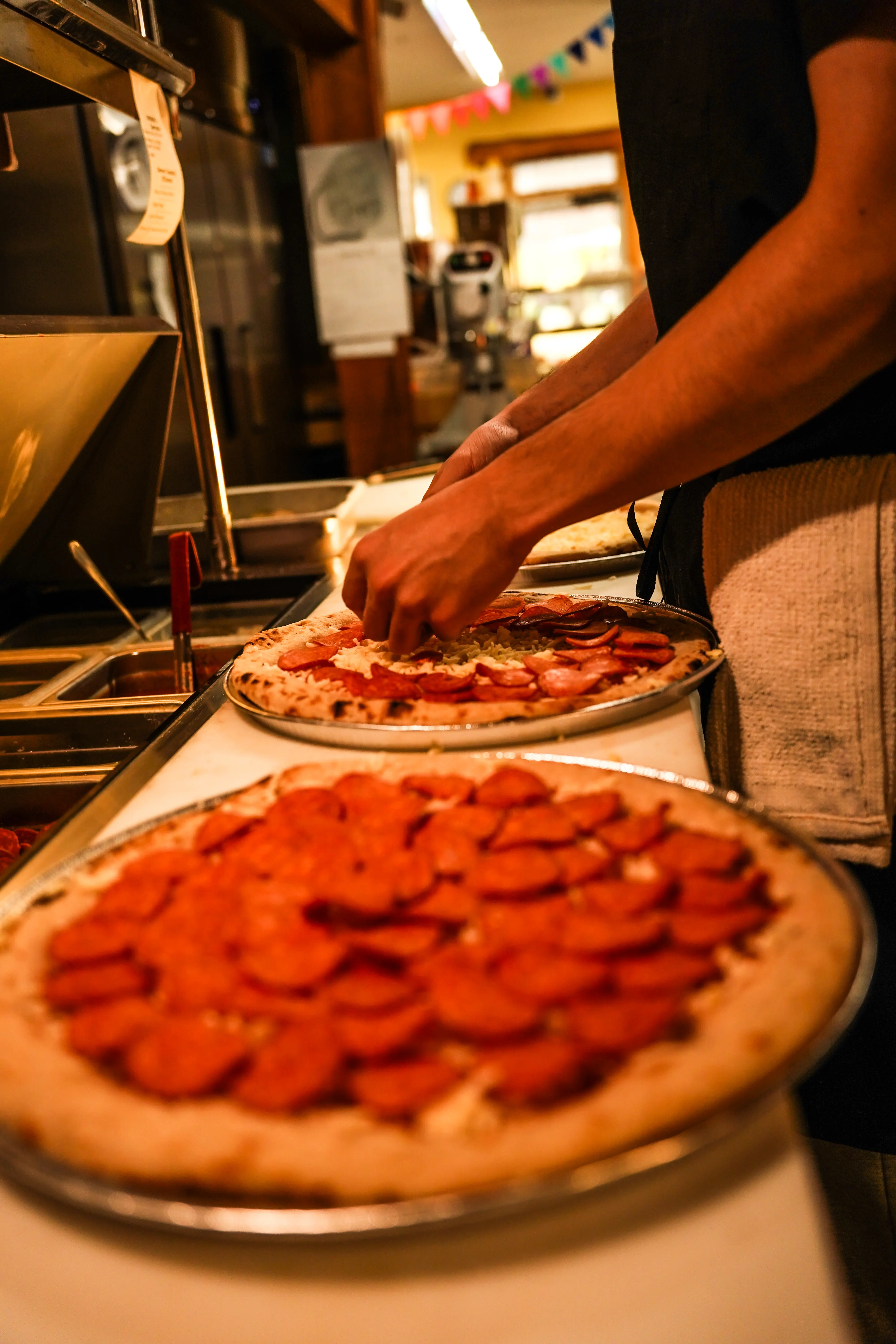 Employee placing pepperoni on a pizza, with a finished pepperoni pizza in the foreground.